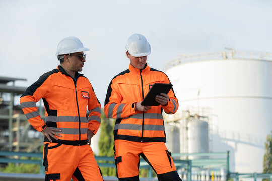 Two industrial workers in orange safety gear and helmets inspect data on a tablet at an oil refinery or chemical plant.
