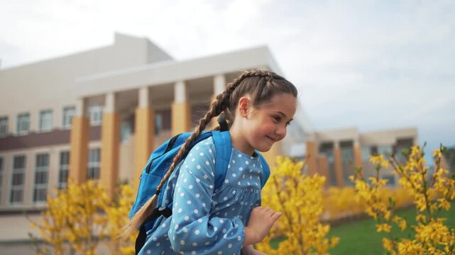 Happy schoolgirl with braids raises hands outside school. Cheerful pupil with backpack smiles near school building. Excited child celebrates. Joyful kid enjoys education. Student laughs outdoors.