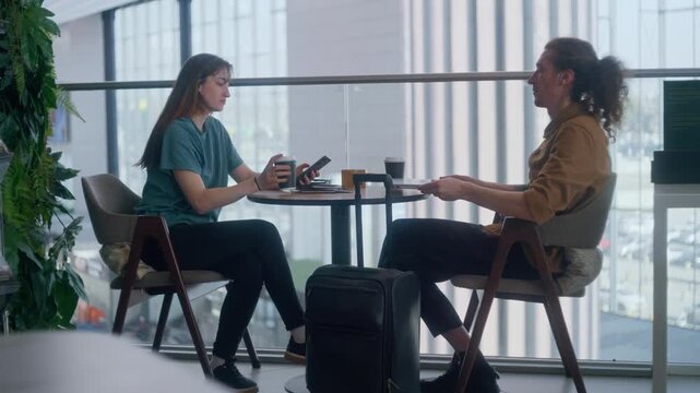 Young man and woman sitting at a table with a laptop, coffee, and luggage in an airport terminal