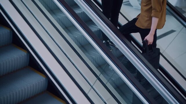 A young man and woman travel together, climbing an airport escalator hand in hand. The clean, contemporary architecture and soft lighting frame their relaxed, united ascent.