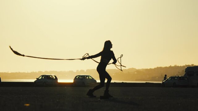 Woman performing rope tricks by the water at sunset with cars in the background