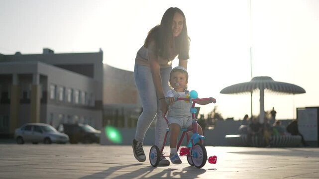 Mother pushes toddler riding tricycle outdoor. Happy mother guides young toddler walking. Sunny summer weather shines brightly outdoor. Mother supports riding child. Tricycle rolls upon square.