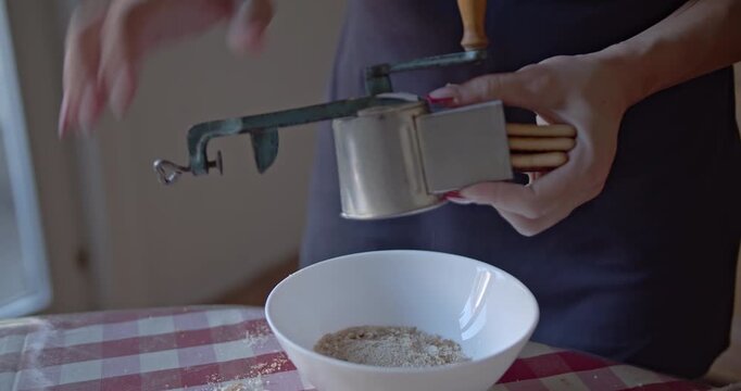 Close-up of a person's hands operating a vintage manual grater to make fresh breadcrumbs into a white bowl. Perfect for illustrating homemade cooking and traditional food preparation.