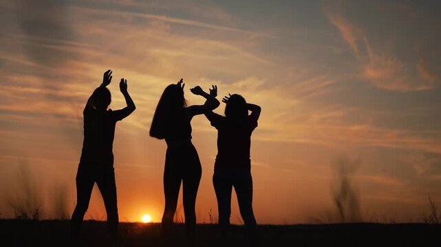 Silhouettes of friends dancing at sunset. Group of girls raise hands in joy under orange sky. Friends celebrate freedom outdoors. Sunset dance party in field. Happy people enjoy dusk together.
