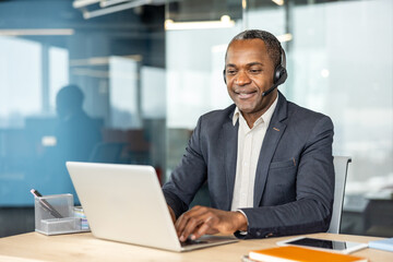 Mature smiling man in a modern office wearing a headset and typing on a laptop, providing customer support and remote communication during a virtual meeting or teleconference