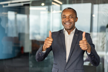 Fototapeta premium Happy professional man in a suit standing in an office, confidently smiling at the camera and holding both thumbs up, symbolizing approval and success