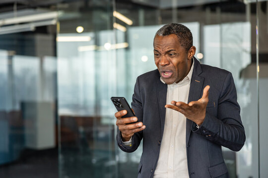 Mature executive showing frustration while browsing unpleasant content on his mobile phone, experiencing stress and annoyance in a modern office environment
