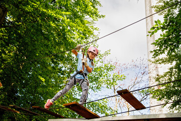 Naklejka premium Happy little girl climbing rope course in forest adventure park, child wearing helmet and harness enjoying outdoor activity, real life moment. School child having fun.