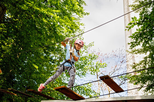 Happy little girl climbing rope course in forest adventure park, child wearing helmet and harness enjoying outdoor activity, real life moment. School child having fun.