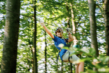 Fototapeta premium Teenage boy climbing rope course in forest adventure park, child wearing helmet and harness enjoying outdoor challenge, real life.