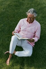 Fototapeta premium Elderly man reading open book seated on grassy ground