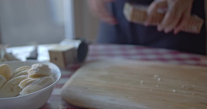 A woman's hands meticulously prepare a sweet banana and biscuit dessert in a home kitchen. She grates biscuits into a bowl, next to sliced bananas, on a charming checkered tablecloth.