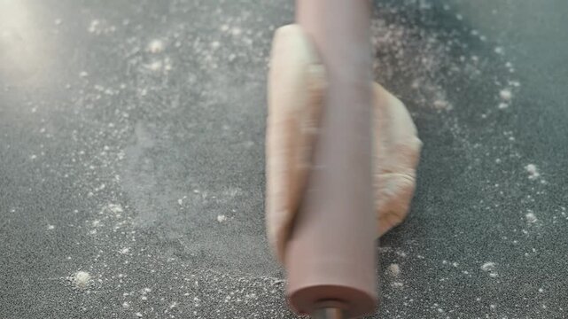 A person rolls out dough on a flat surface using a rolling pin in a kitchen. The action occurs indoors and involves preparing the dough for baking, showcasing the process of making bread or pastry