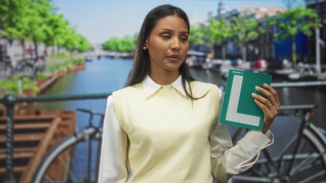 Woman holding green learner l plate with right hand on canal street by bicycle and railing; uncertainty learning progress.