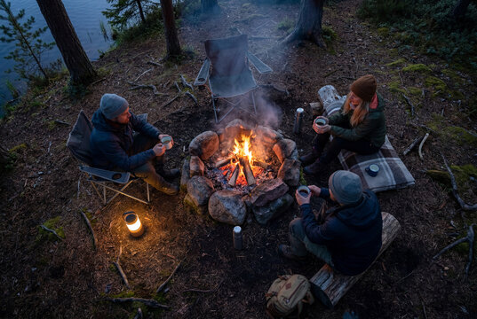 Three adults gather around a campfire by the lake at dusk, sharing hot drinks in a digital detox camping moment.