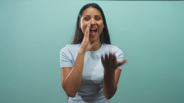 Woman, young brunette, cupping hand to mouth and showing ring while smiling against a teal studio wall in mid call gesture; playful energy.