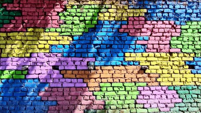 A vibrant brick wall painted in a patchwork of bright colors�blue, pink, yellow, green, and purple�forms a textured mosaic, with sunlight casting subtle shadows across the uneven surface.