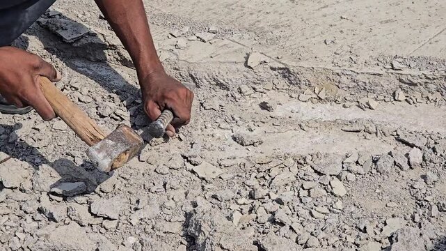 A construction worker manually demolishes a concrete slab using a sledgehammer and a chisel. The surrounding area is cluttered with fragments of rock, gray rubble, and gravel from the broken pavement.