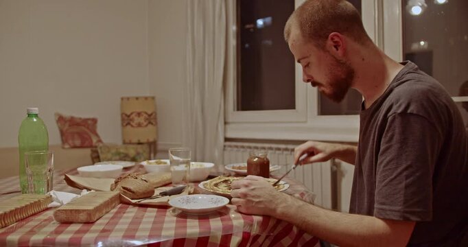 A young man enjoys a sweet treat, carefully spreading rich chocolate hazelnut paste onto a warm pancake at his dining table, creating a cozy and comforting moment at home.