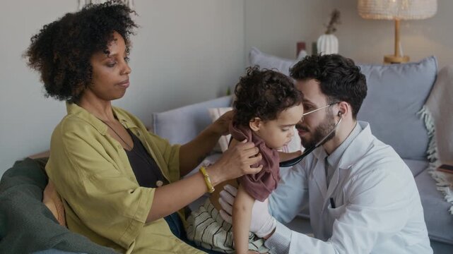 Medium shot of male multiethnic doctor wearing white coat listening to breath of African American toddler sitting with mother during visit at home