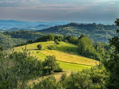 Aerial view of the rolling Tortona Hills with lush green meadows, dense forests, and a small hilltop village under a soft evening sky in Tortona, Piedmont, Italy.