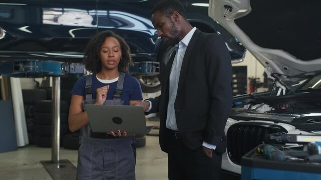 Female mechanic in overalls showing a client, interested African American businessman, a vehicle diagnostic on a laptop in a car repair shop.