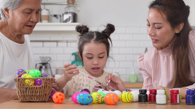 Asian little girl painting colorful easter eggs with brush while grandmother helping her at kitchen table, Three generations family spring holiday celebration, creative art craft learning, easter day