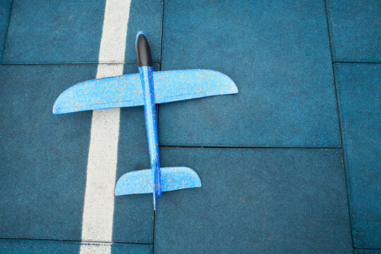 Wide urban court view with plane on surface, blue speckled glider resting on rubber tiles, apartment blocks and street visible in background, community