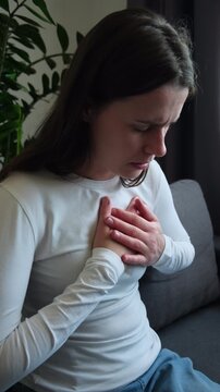 Close up of suffering acute cardiac young caucasian woman hand touching on her chest pain, female breathing problem, pressure sitting alone on couch at home. Heart attack, health care concept