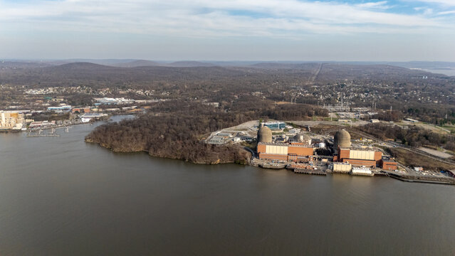 Aerial view of the Indian Point Energy Center nuclear power plant facility on the Hudson River bank surrounded by dense forests and rolling hills in Buchanan, New York, United States.