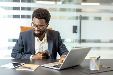 Professional african ethnic man experiencing abdominal pain while working on a laptop at a...