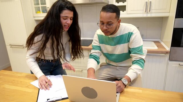 Multiracial young couple working on their home finances and budget, using a laptop and documents on the kitchen counter, showing a mix of concentration, discussion, and finally satisfaction