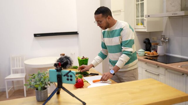 Young male food blogger recording a video for his channel, talking to the camera and demonstrating how to peel vegetables in a modern kitchen while explaining a healthy recipe