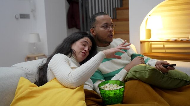 Happy couple watching a movie together on the sofa at night, eating popcorn and enjoying their leisure time at home. The man uses the remote control while the woman cuddles up to him