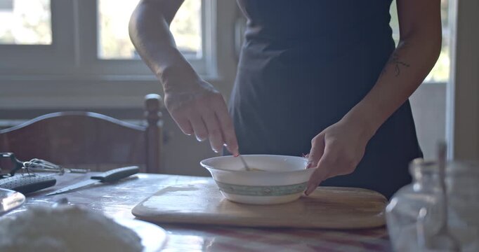 A close-up captures a woman's hands expertly mixing ingredients in a ceramic bowl on a rustic kitchen table.