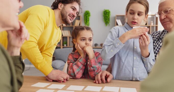 Family children parents play boardgame with cards. Kids focus at the table at home, taking turns. Friendly smiles show cooperation and memory practice indoors. Clear idea of family learning.