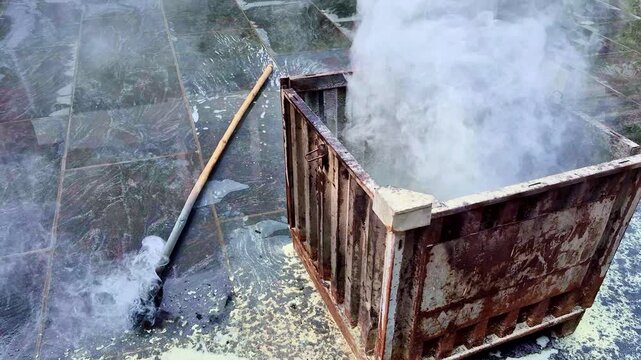 A rusted metal container sits on a tiled ground, emitting thick white smoke after being doused, while a long wooden stick with a charred end lies nearby, still smoldering extinguished fire.