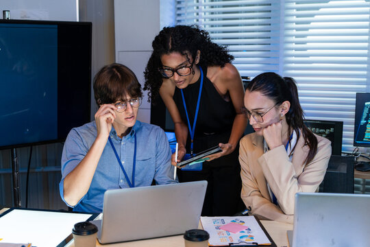 Serious multiracial tech team focused on laptop screen while working in office. Young professional colleagues using digital tablet and computers for software development during group project meeting