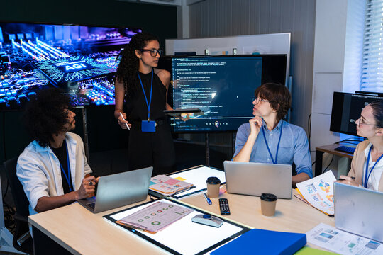 Professional female leader presents artificial intelligence and software code to diverse tech team in office. Young experts using laptops to develop digital solutions during corporate meeting