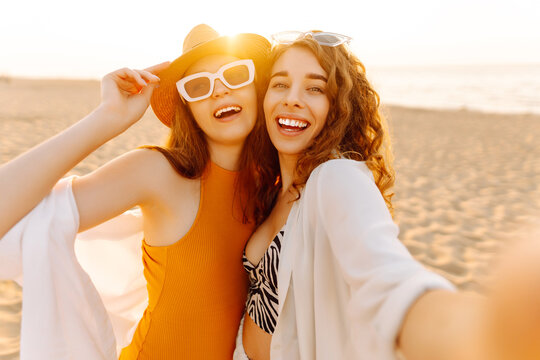 Two girlfriends take selfies on a phone on a sunny beach. Young women enjoy a beach vacation, blogging at sunset. Concepts of relaxation, friendship, and fun.
