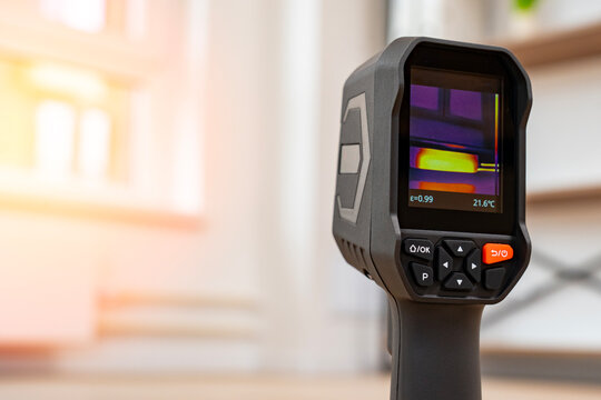 Thermal imaging camera displays heat patterns on its screen. Color gradients show temperature variations clearly. The device is positioned indoors with soft background lighting
