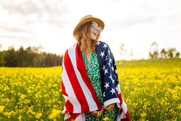 Woman stands in field of yellow flowers, waving American flag. Beautiful woman enjoys the blooming field with an American flag in her hands. Concepts of independence, relaxation, and beauty. © maxbelchenko