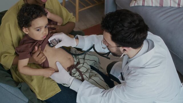 View from above of male multiethnic paediatrician in white coat using stethoscope while examining small patients breath sitting with mom during home appointment