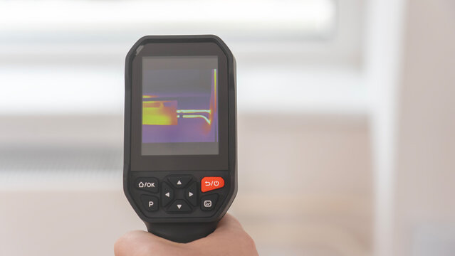 Person holds a black thermal camera. The screen shows color-coded heat patterns. Yellow shows warmth, red indicates heat, purple reflects cooler areas. This tool identifies temperature variations