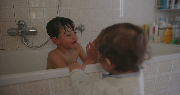 Older brother smiling in bathtub while baby toddler reaches to interact showing playful sibling bond joy and family connection during everyday bath time at home
