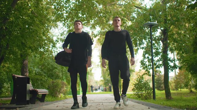 two men walking through leafy park carrying duffel bag, casual athletic clothing, confident stride past bench and lamppost with sunlight filtering through trees, relaxed friendship vibe as partners