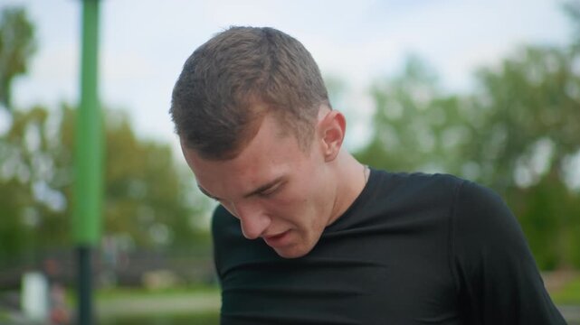 white athlete catching breath outdoors postsprint pause on park pullup bar, heavy breathing, sweat on brow, short cropped hair, black training shirt, blurred leafy background, natural light,