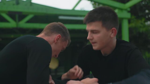 two white men grappling under green pavilion intense outdoor sparring with aggressive clinch exchanges and takedown attempts strained faces sweatstreaked black shirts urban park structure