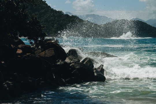 Ondas colidindo com rochas escuras, criando um spray de &aacute;gua dram&aacute;tico sob o sol. Ao fundo, montanhas cobertas por floresta tropical e c&eacute;u azul.