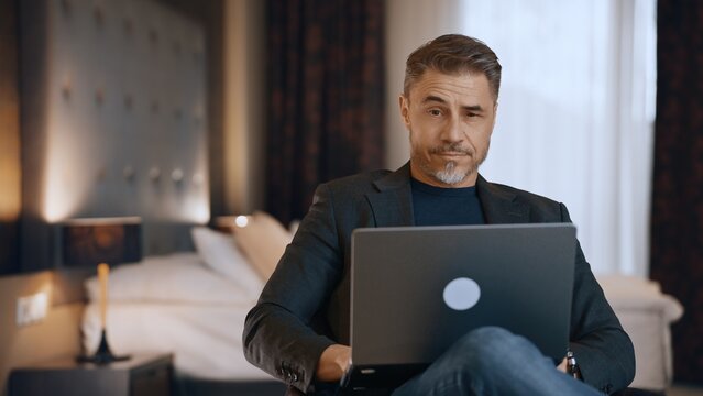 Confident mature male professional using laptop in luxurious hotel room. Businessman working at home with computer, thinking. Portrait of serious mid adult man. Entrepreneur managing business online.